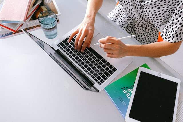 a-person-typing-on-a-laptop-with-a-stylus-in-hand-surrounded-by-books-a-glass-of-water-and-a-tablet-on-a-desk