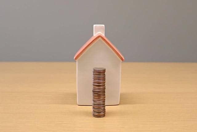 stack-of-coins-in-front-of-a-small-ceramic-house-on-a-wooden-surface-symbolizing-property-value-or-home-investment